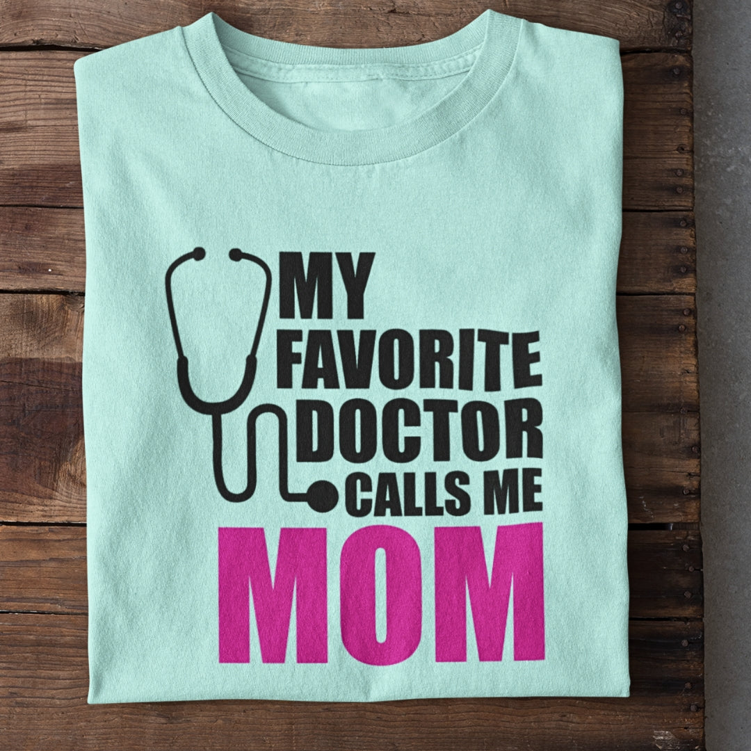 Light blue folded t-shirt on wooden surface with text and stethoscope graphic saying, 'My favorite doctor calls me mom'