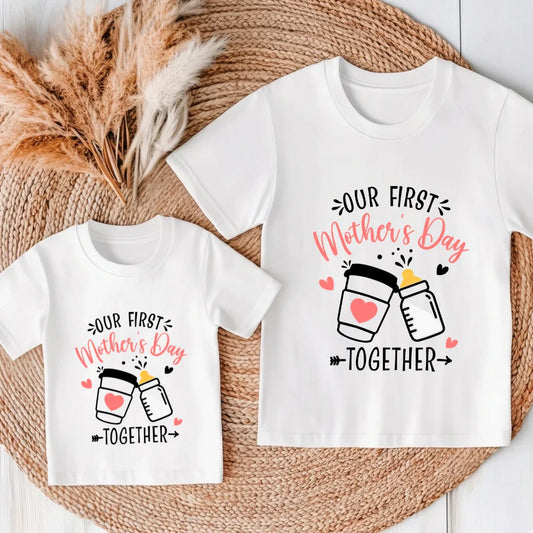 Two white t-shirts with 'Our First Mother's Day Together' text and jar design on a woven surface.